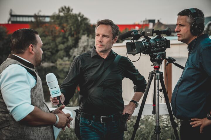A man operates a camera while interviewing a woman outdoors.